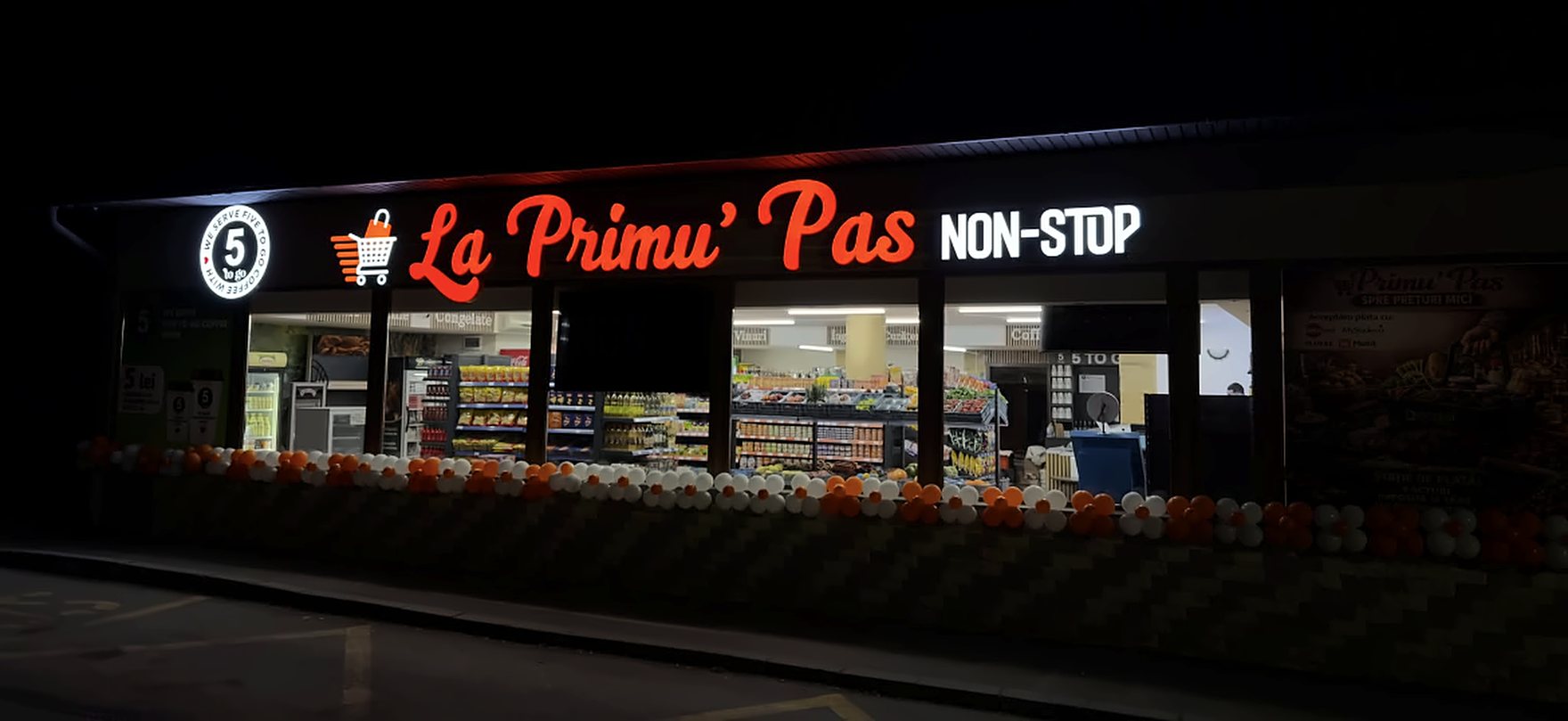 Magazinul La Primu' Pas Non-Stop seara: firma roșie aprinsă, raioanele luminate și baloane portocalii la vitrină